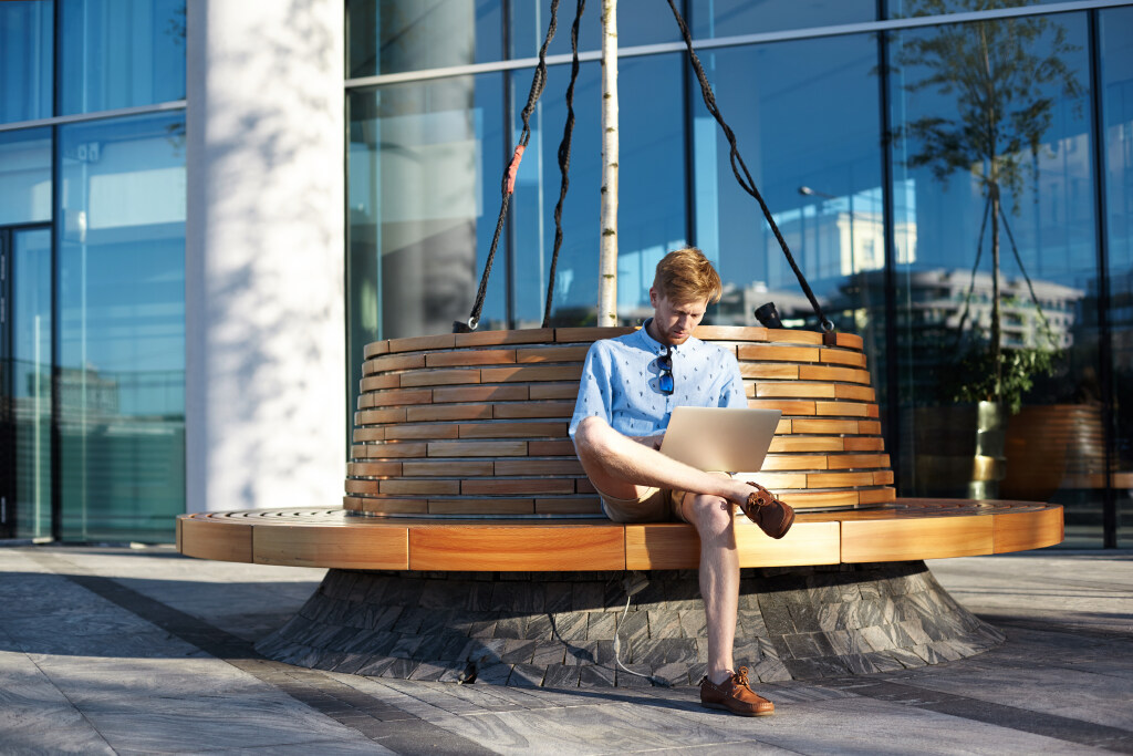Outdoor picture of freelance worker using a laptop computer for remote work, sitting on bench in urban surroundings, enjoying free public wifi